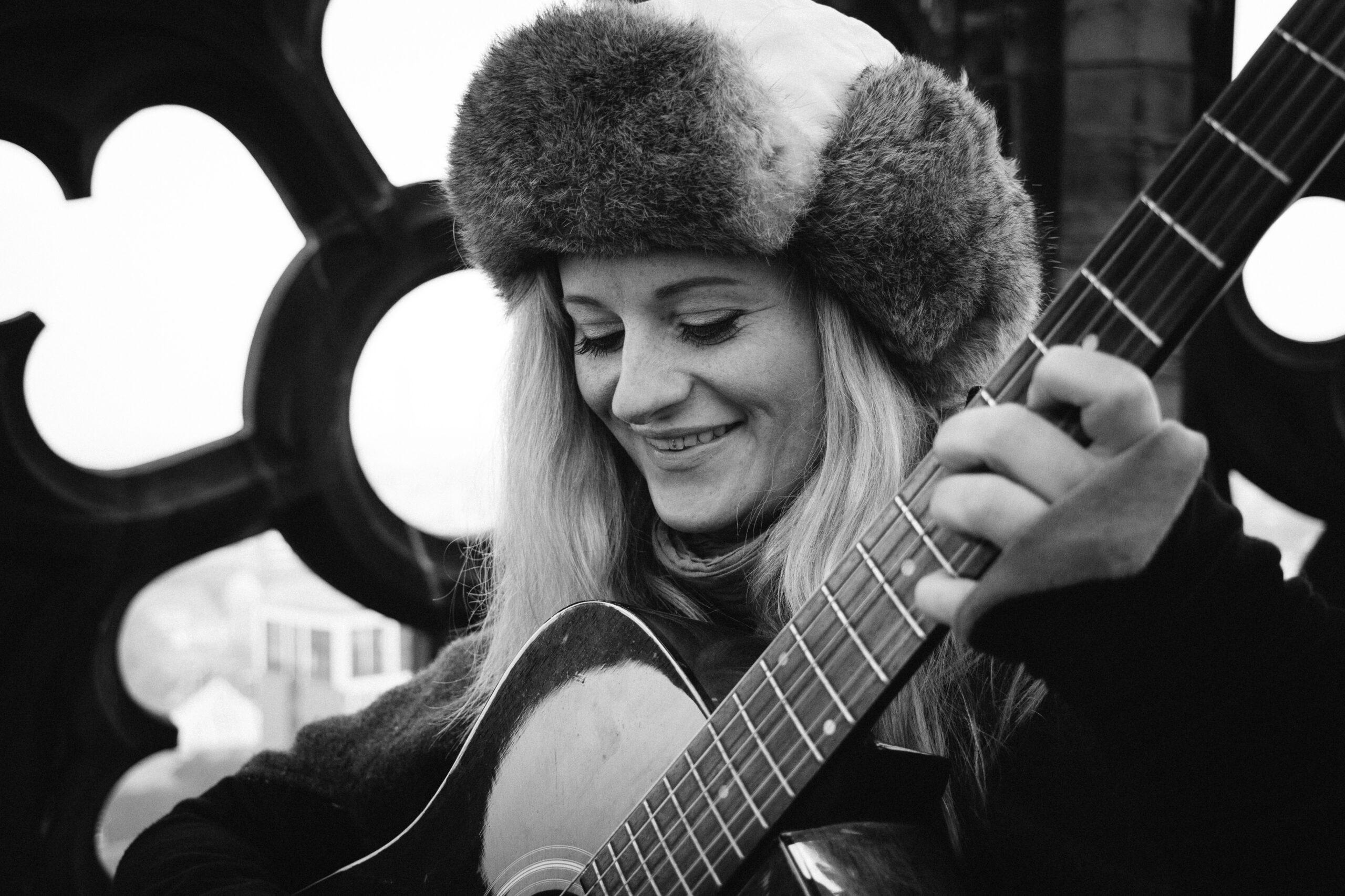 Auf dem Bild ist Marta Latour, die Türmerin von Münster, zu sehen. Sie trägt eine auffällige Fellmütze, lächelt und greift einen Akkord auf einer Gitarre. Im Hintergrund Maßwerk der Lambertikirche.